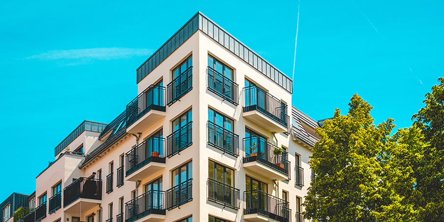 A white multi-story apartment building, camera angled up to capture a bright blue sky in the background
