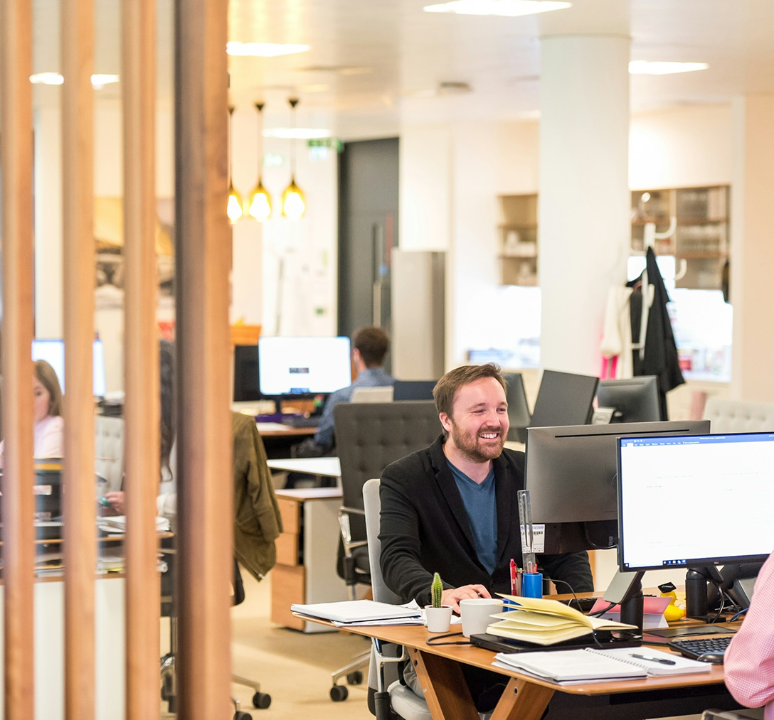 man working at his computer smiling in an office where other people are working